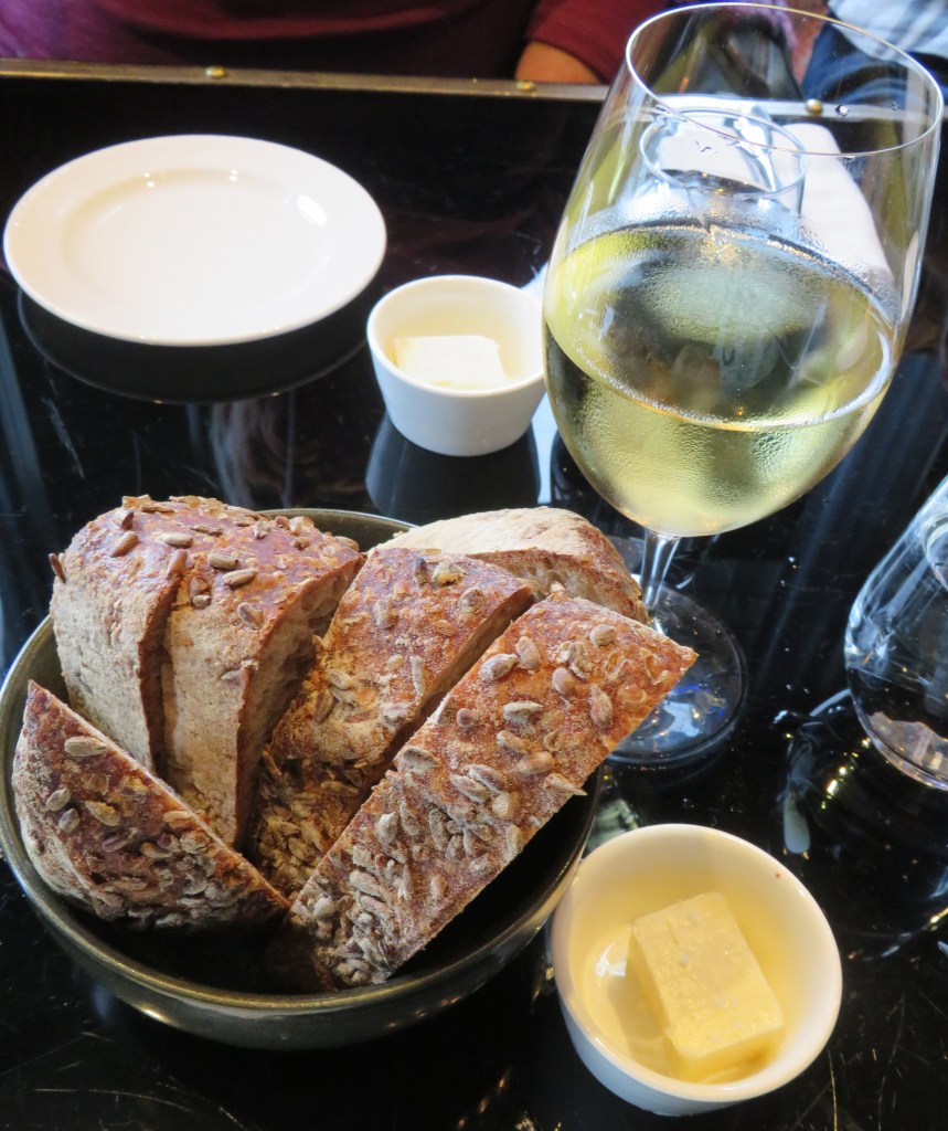 A bowl of sliced artisanal bread with sunflower seeds, accompanied by a small dish of butter and a glass of white wine on a black table.